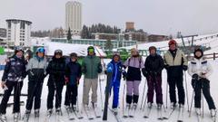 10 skiers lined up in a row. The sixth skier is the instructor Luca, who has his right ski raised.