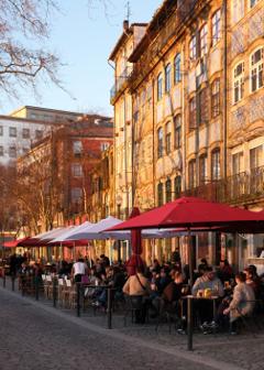 People socialise under red and white canopies. The buildings behind them are golden, with the shadows of nearby trees imparted on their beautifully decorated facades.