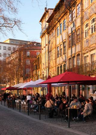 People socialise under red and white canopies. The buildings behind them are golden, with the shadows of nearby trees imparted on their beautifully decorated facades.
