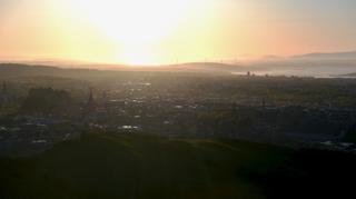 A hazy cityscape in soft, diffused lighting of a late evening. In the far distance, rivers and hillside.