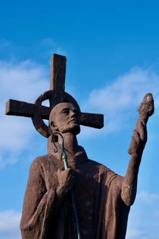 A statue of a saintly figure holding up a cross with one hand while raising his other hand in blessing or pointing gesture.
