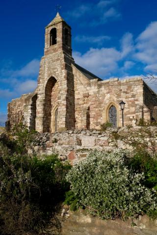 An old stone building with a bell tower surrounded by greenery and clear skies.
