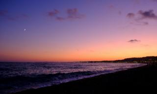 The pebbled beach in Nice at dusk under a purple-orange sky with a crescent moon and city lights along the coast.