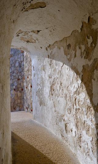 A sunlit, peeling whitewashed passageway leading to a stone wall.