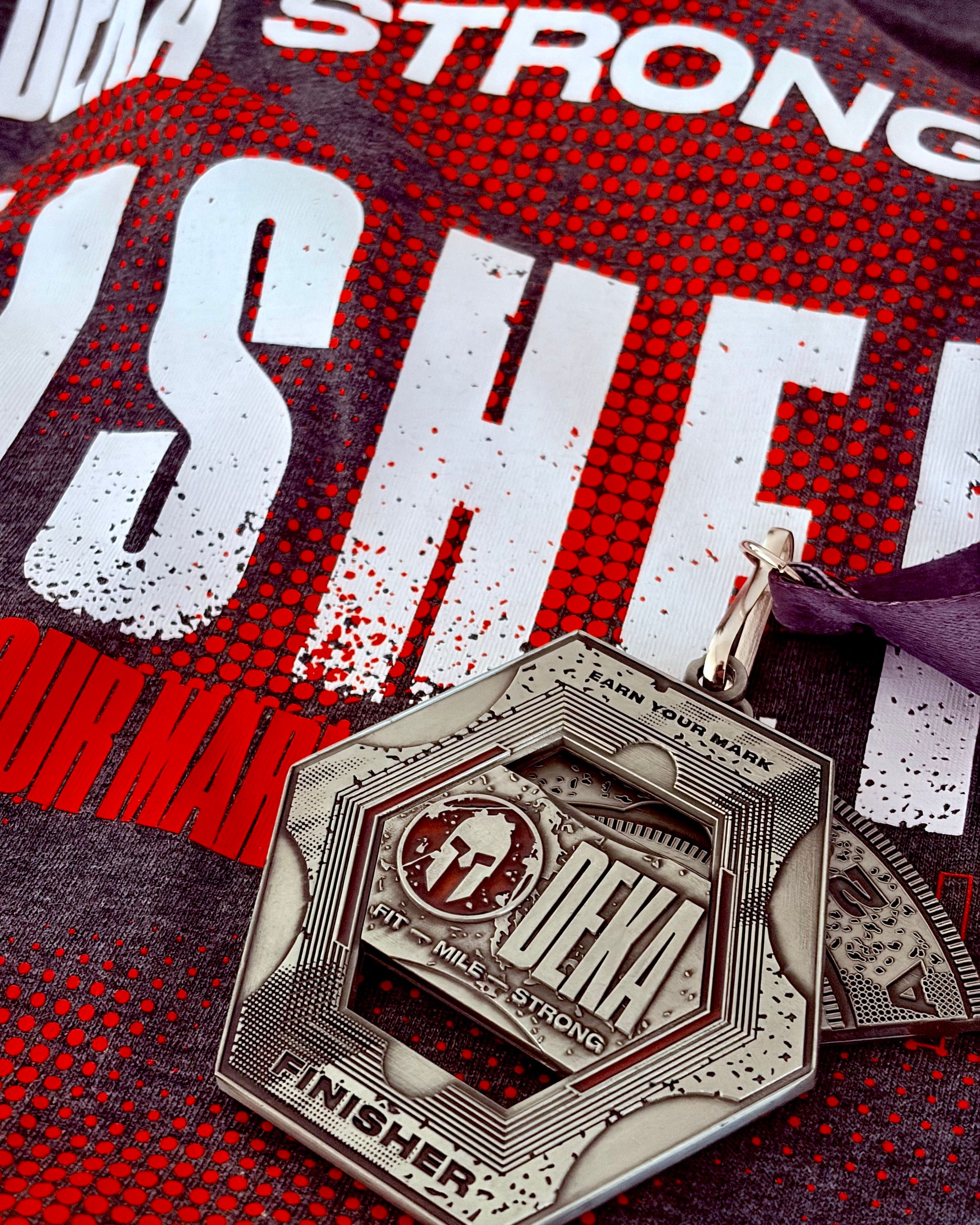 Dark grey hexagonal finisher’s medal sat on a grey and red finisher’s t-shirt.